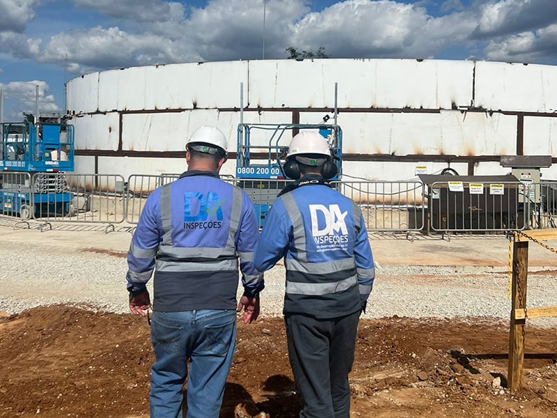 Dois técnicos da D&A Inspeções de costas, observando um grande tanque de armazenamento ao ar livre. Eles usam uniformes com o logo da D&A visível nas costas e capacetes de segurança.